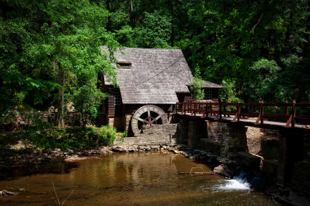 A serene forest scene featuring a watermill building beside a flowing stream, connected by a wooden bridge, captured in high-definition quality.