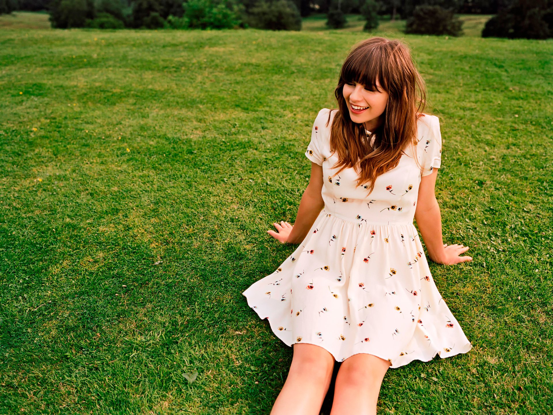 Gabrielle Aplin, English singer with brunette hair, smiles while sitting on grass in a white dress, captured in a bright HD desktop wallpaper.