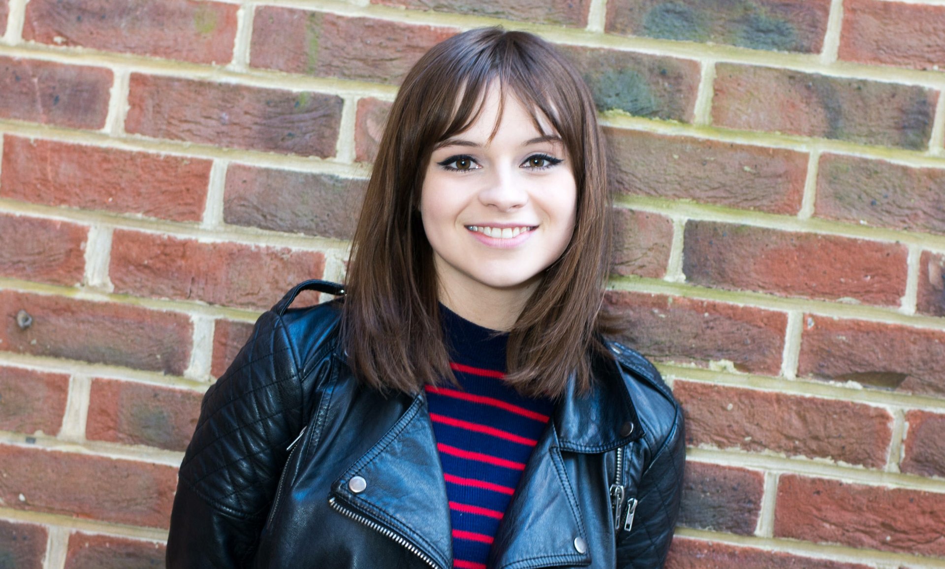 HD desktop wallpaper featuring English singer Gabrielle Aplin with brunette hair, brown eyes, and a warm smile against a brick wall background.