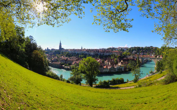 5K Ultra HD panorama of Bern, Switzerland: riverside cityscape with the Aare River curving around historic buildings and bridges, framed by a grassy hillside and leafy trees.