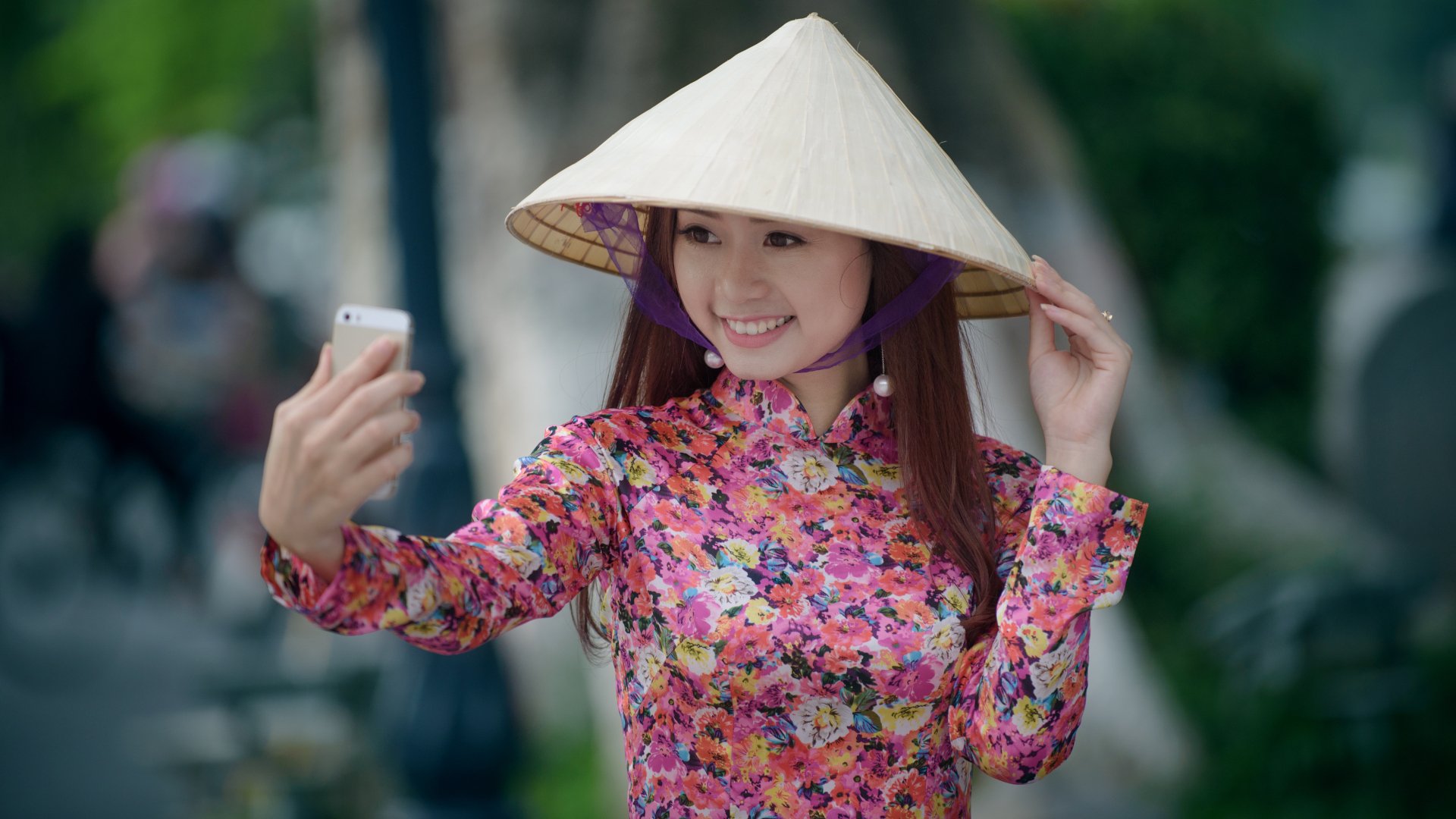 Asian woman with brunette hair smiling and wearing a traditional costume and conical hat, taking a selfie; 4K Ultra HD PC desktop wallpaper and background.