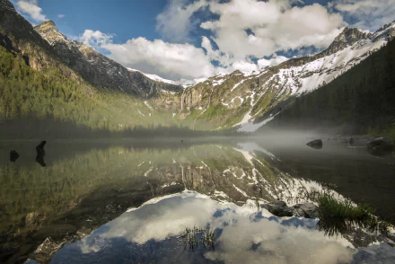 A serene mountain landscape in Glacier National Park, Montana, featuring a tranquil lake reflecting towering peaks and lush greenery under a cloudy sky.