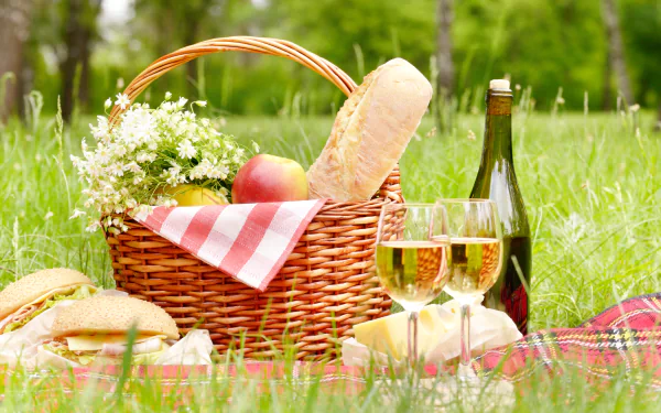 A 4K Ultra HD still life desktop wallpaper of a picnic scene with a wicker basket, bread, sandwiches, wine bottle, two glasses of wine, and fresh grass.