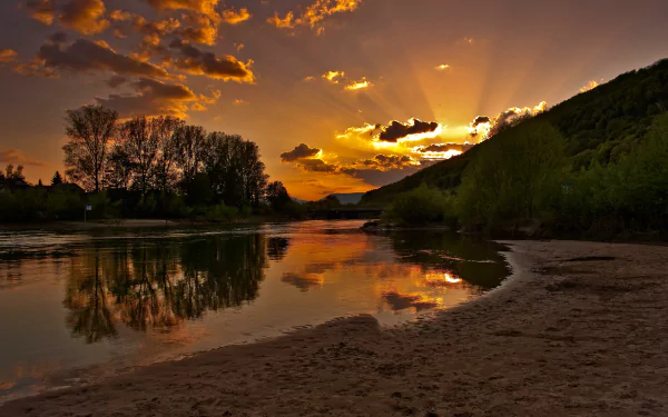4K Ultra HD wallpaper of a serene river at sunset, with vibrant orange skies reflecting on calm water, framed by trees and hills in a peaceful natural setting.
