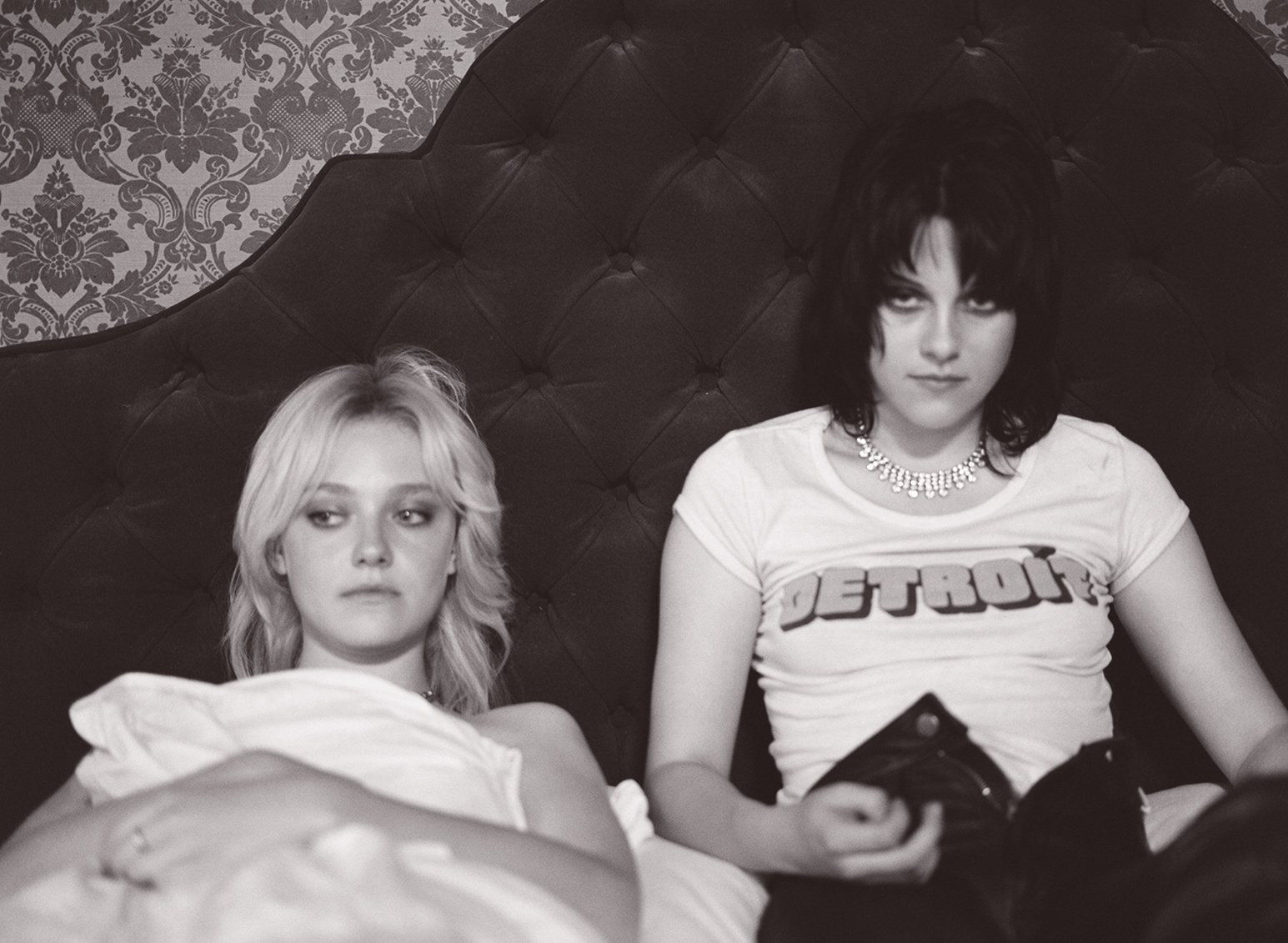 Black-and-white HD desktop wallpaper featuring Kristen Stewart and Dakota Fanning from the movie The Runaways, sitting on a dark tufted headboard.