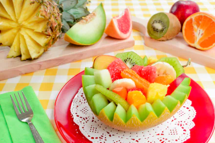 A vibrant 4K Ultra HD desktop wallpaper showcasing a colorful assortment of melon, orange, kiwi, and pineapple fruit artfully arranged on a red plate over a yellow checkered tablecloth.