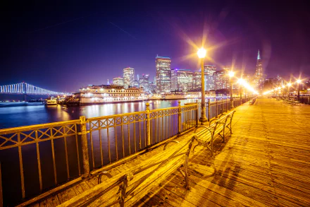 Night view of San Francisco's Pier 7, featuring benches and lamp posts along the waterfront, with the Bay Bridge illuminating the city skyline in the background.