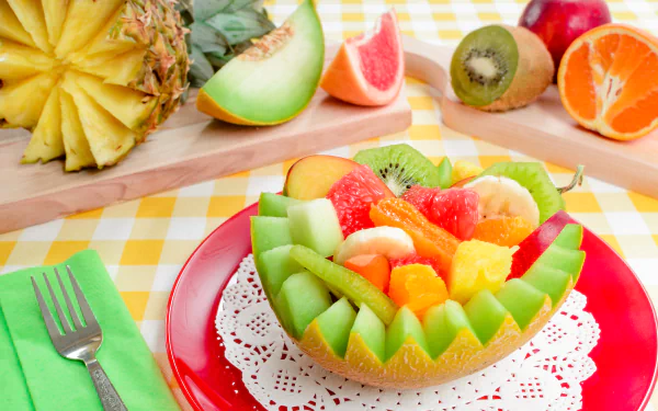 A vibrant 4K Ultra HD desktop wallpaper showcasing a colorful assortment of melon, orange, kiwi, and pineapple fruit artfully arranged on a red plate over a yellow checkered tablecloth.