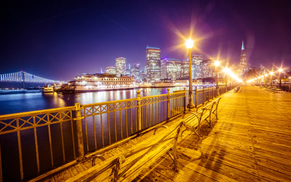 Night view of San Francisco's Pier 7, featuring benches and lamp posts along the waterfront, with the Bay Bridge illuminating the city skyline in the background.