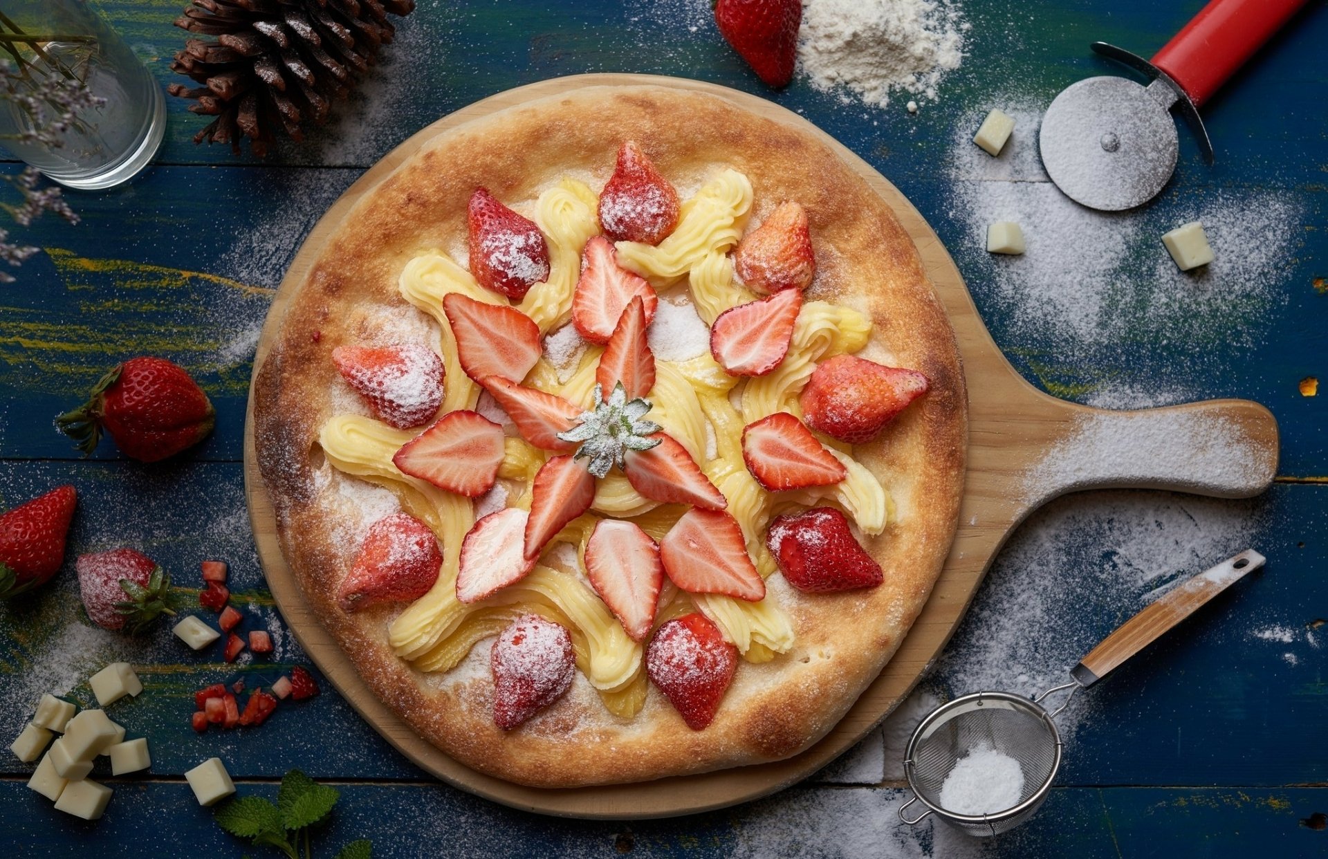 HD desktop wallpaper featuring a still life of a strawberry and cream cake on a wooden board, surrounded by flour, a glass, a pine cone, and baking utensils on a blue surface.