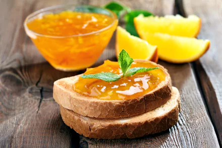 A close-up of two slices of bread topped with orange jam, garnished with mint, beside a bowl of jam and fresh orange wedges, set against a rustic wooden background.