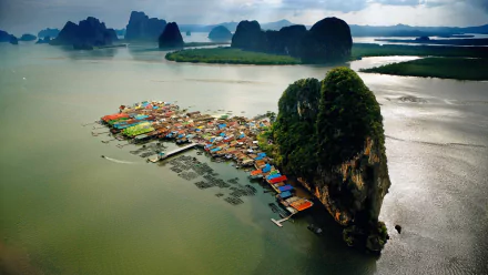 Aerial view of a vibrant man-made village on an island in Phuket, Thailand, surrounded by lush mountains and calm waters, showcasing the region's unique beauty.