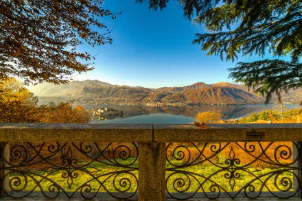Scenic fall view of Lake Orta, Italy, with colorful foliage reflected in the calm water, framed by trees and an ornate stone railing in this HD desktop wallpaper.