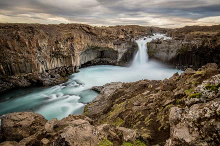 A serene view of Aldeyjarfoss, featuring a majestic waterfall cascading over cliffs into a tranquil river, set against a dramatic natural landscape. Perfect HD wallpaper for nature lovers.