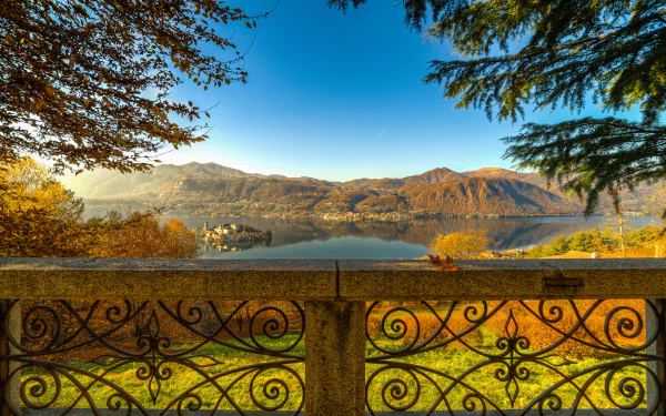 Scenic fall view of Lake Orta, Italy, with colorful foliage reflected in the calm water, framed by trees and an ornate stone railing in this HD desktop wallpaper.