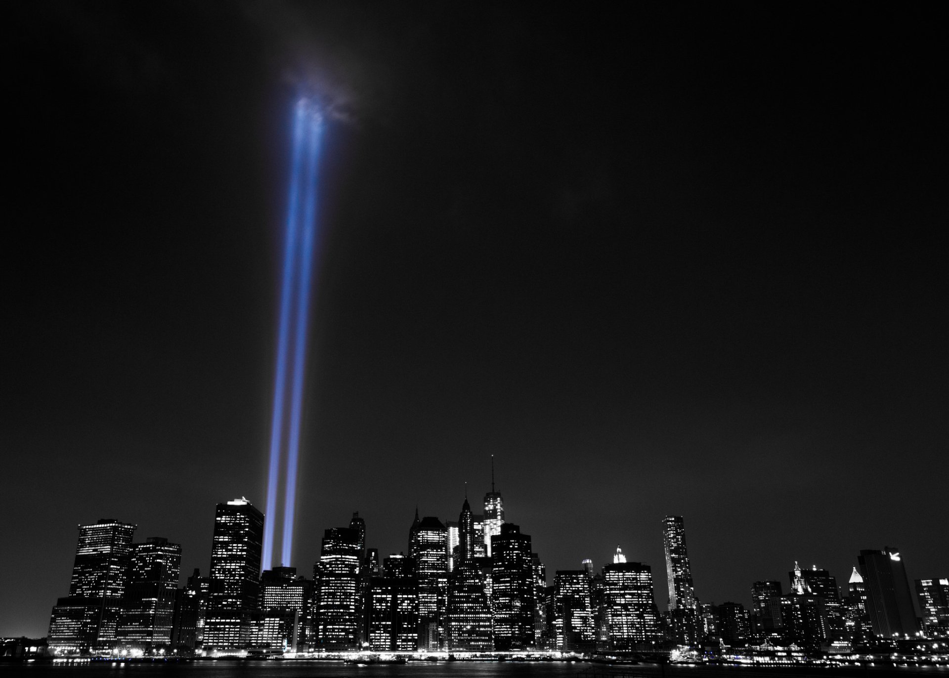 Selective color image of New York City skyline at night with two bright light beams shining upward from skyscrapers, highlighting the man-made urban landscape.