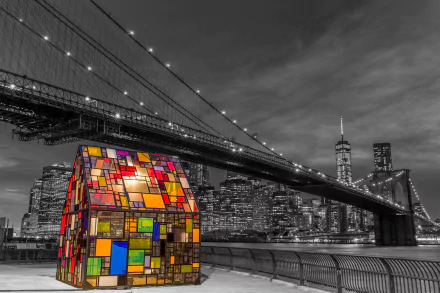 Night view of New York City featuring the illuminated Brooklyn Bridge and a colorful, stained-glass-like structure along the waterfront in this HD cityscape wallpaper.