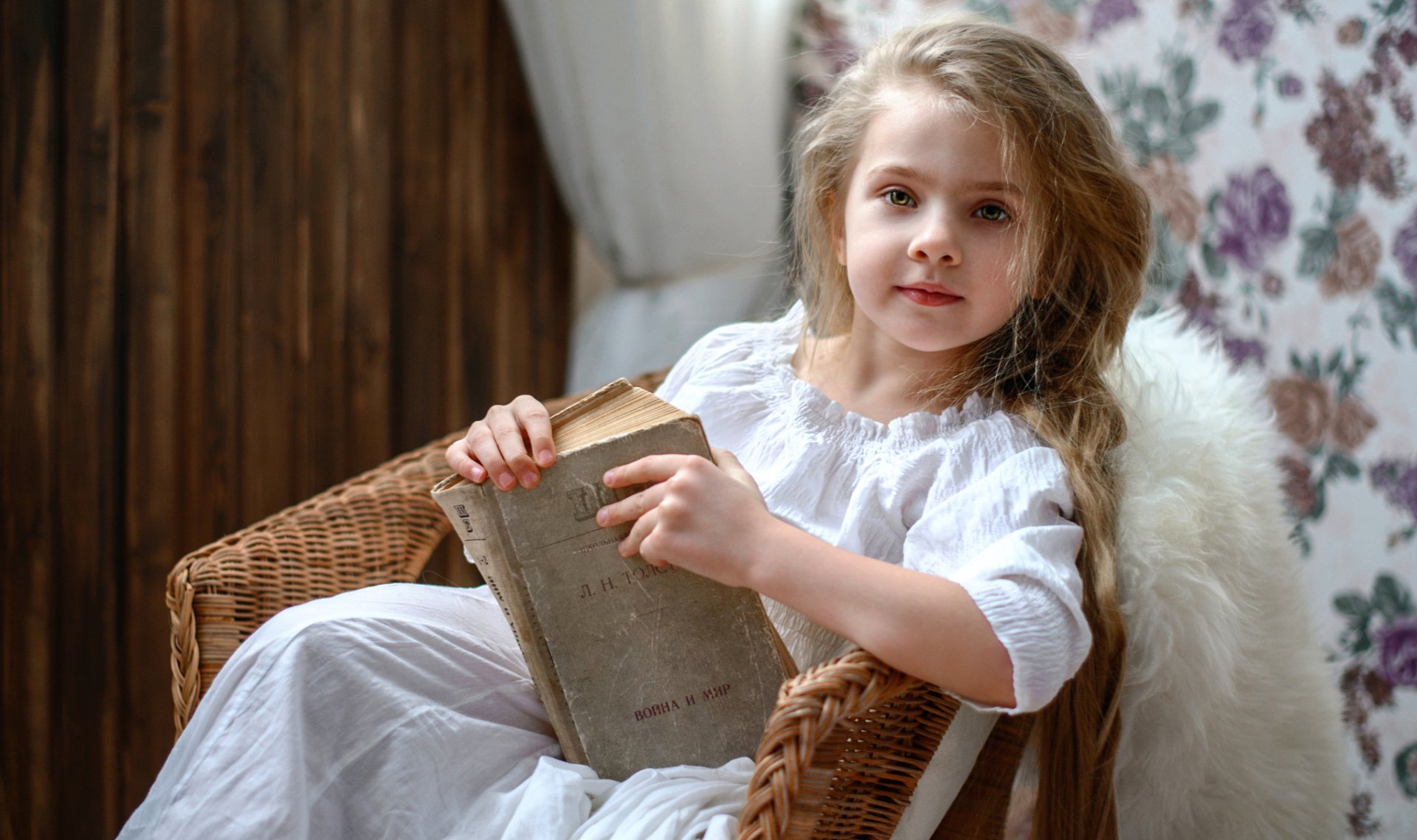 A little girl with long blonde hair and green eyes sits in a wicker chair, holding an old book, against a softly patterned floral background.
