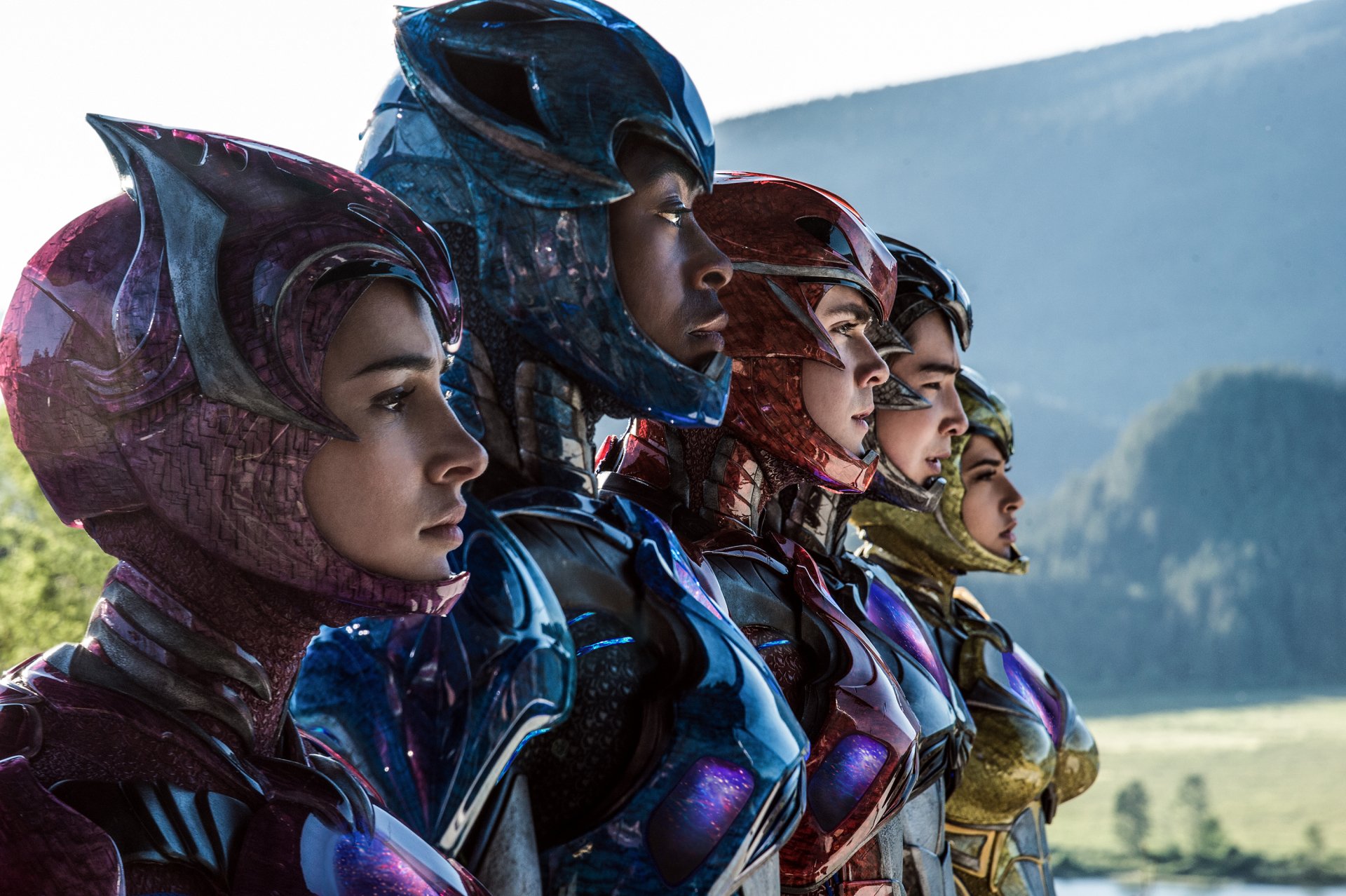 The image features the Power Rangers from the 2017 movie, showcasing the team in colorful suits standing side by side against a natural landscape.