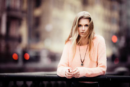 Blonde woman model with brown eyes leans on a railing against a blurred city background — HD PC desktop wallpaper/background.