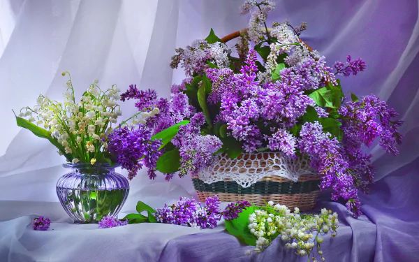A still life HD desktop wallpaper featuring a basket and vase filled with purple lilacs and white lily of the valley flowers, set against soft draped fabric.