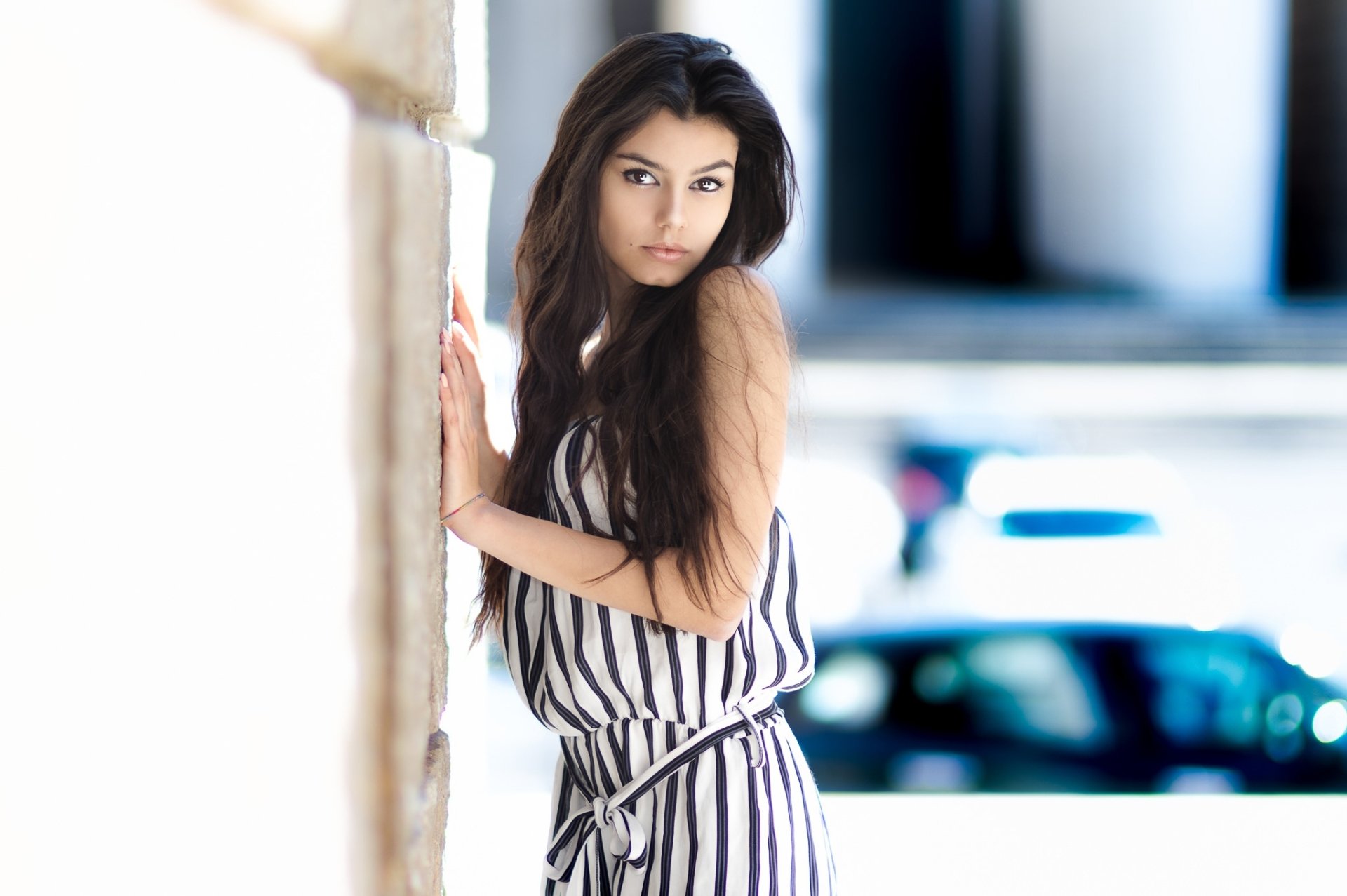 HD desktop wallpaper featuring a brunette woman with brown eyes in a striped outfit, leaning against a stone wall with a blurred background.