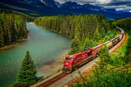A vibrant locomotive traverses a scenic landscape, flanked by a winding river and lush forest, set against majestic mountains in this stunning HD wallpaper.