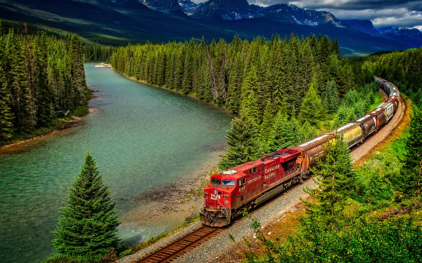 A vibrant locomotive traverses a scenic landscape, flanked by a winding river and lush forest, set against majestic mountains in this stunning HD wallpaper.