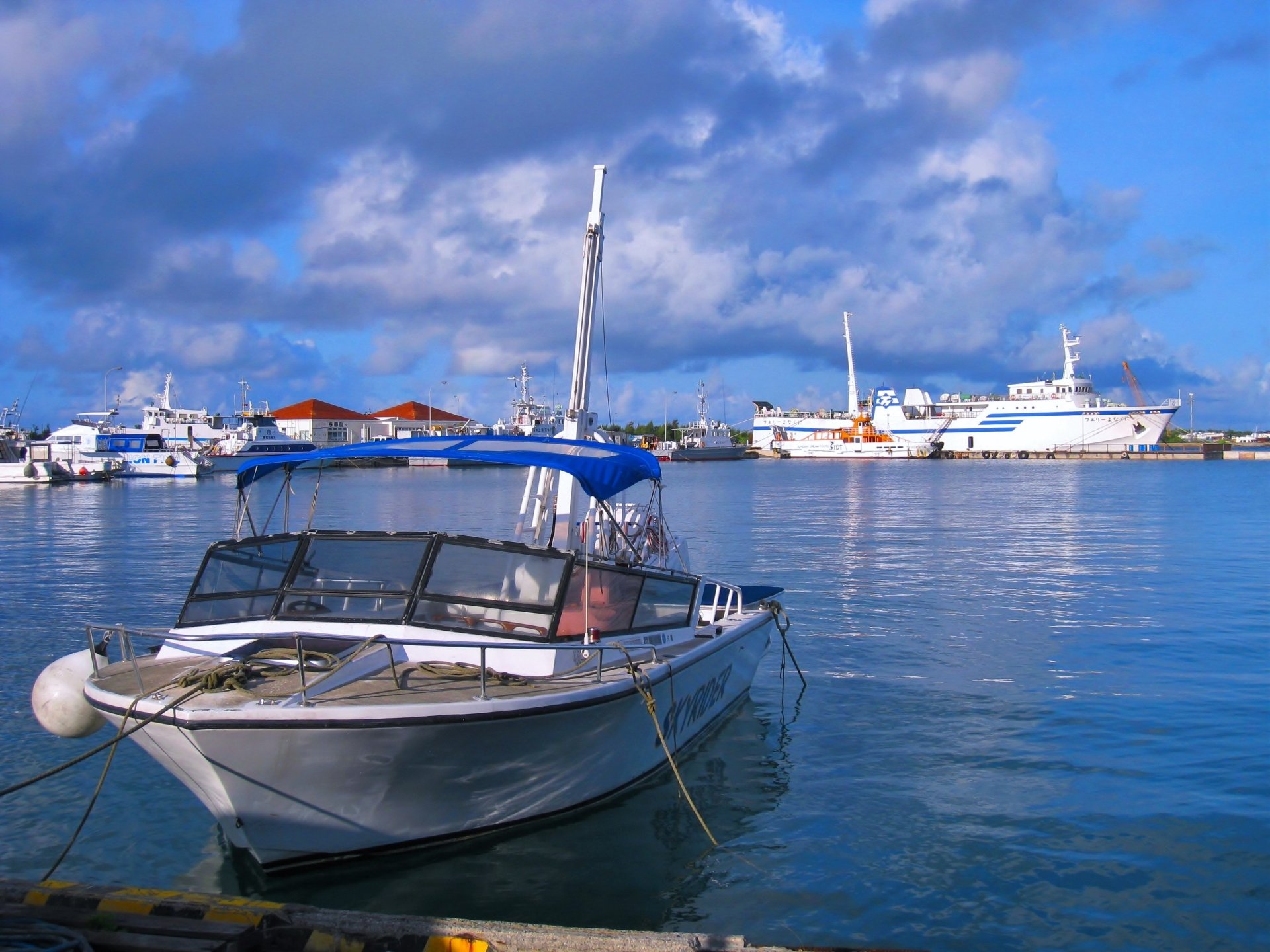 2K Quad HD PC desktop wallpaper of a small motorboat moored in a calm harbor with larger ships and buildings under a blue, cloudy sky — ship, vehicle, boat