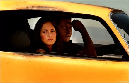 A high-definition wallpaper featuring two characters inside an orange car, set against a dramatic backdrop, inspired by the Transformers movie.