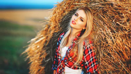 Blonde woman in a red plaid shirt leans against a haystack in a picturesque rural setting. The HD wallpaper captures her casual, relaxed pose.