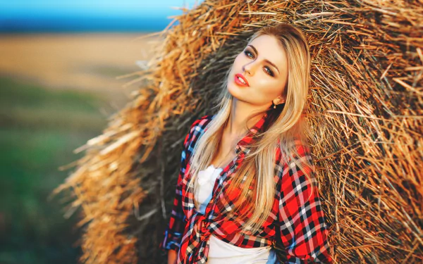 Blonde woman in a red plaid shirt leans against a haystack in a picturesque rural setting. The HD wallpaper captures her casual, relaxed pose.