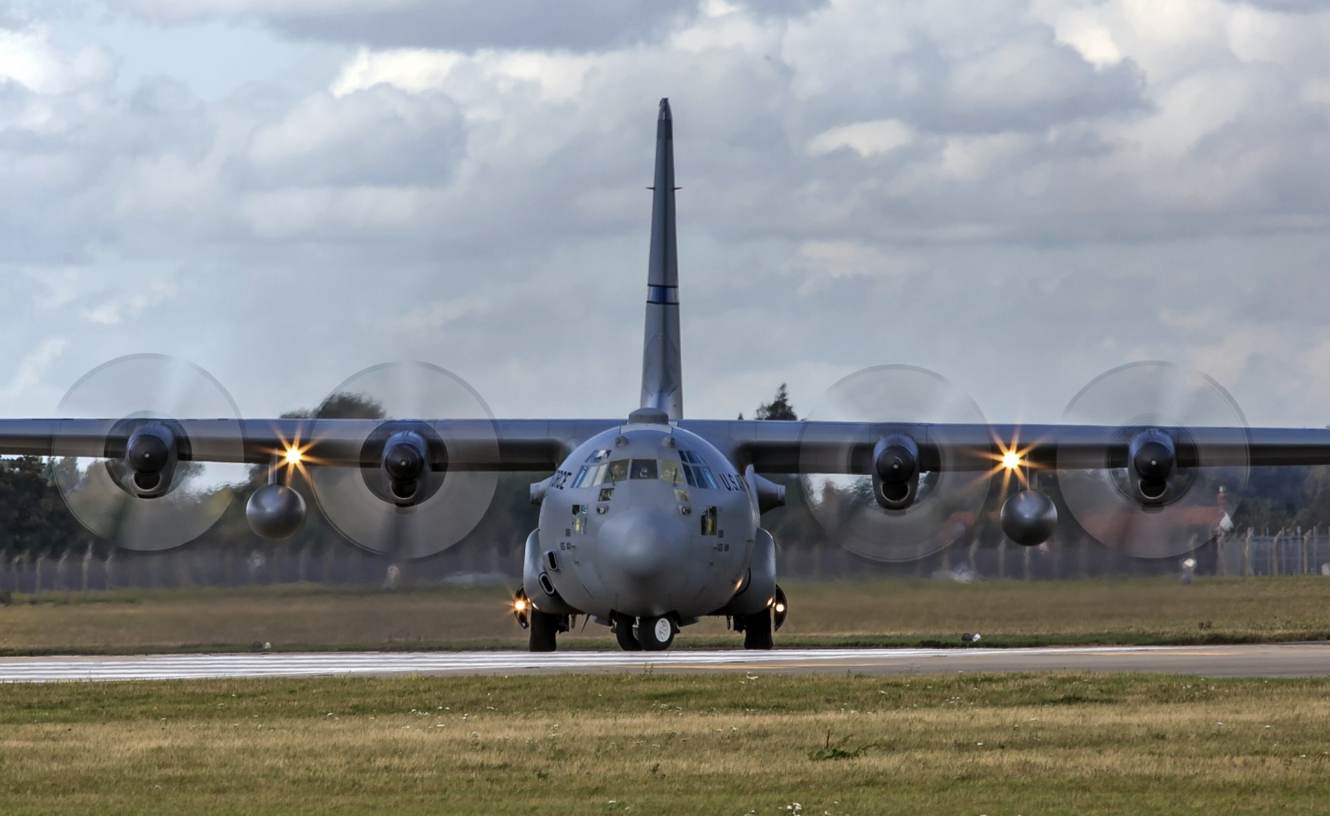 Front view of a Lockheed C-130 Hercules warplane transport aircraft, propellers spinning as it taxis on the runway beneath a cloudy sky — 2K Quad HD PC desktop wallpaper.