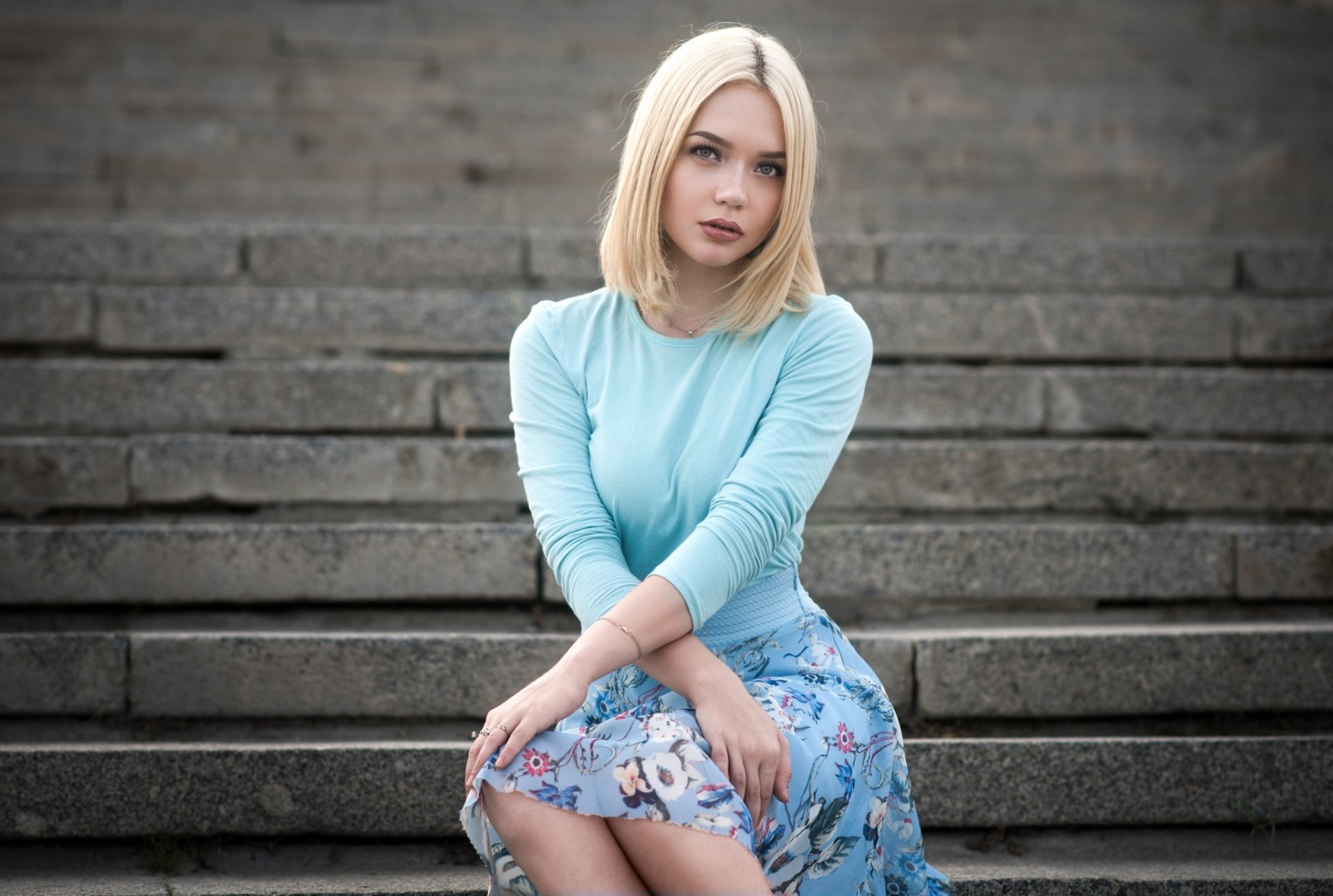 A blonde woman in a blue top and floral skirt sits on stone steps. HD desktop wallpaper and background.