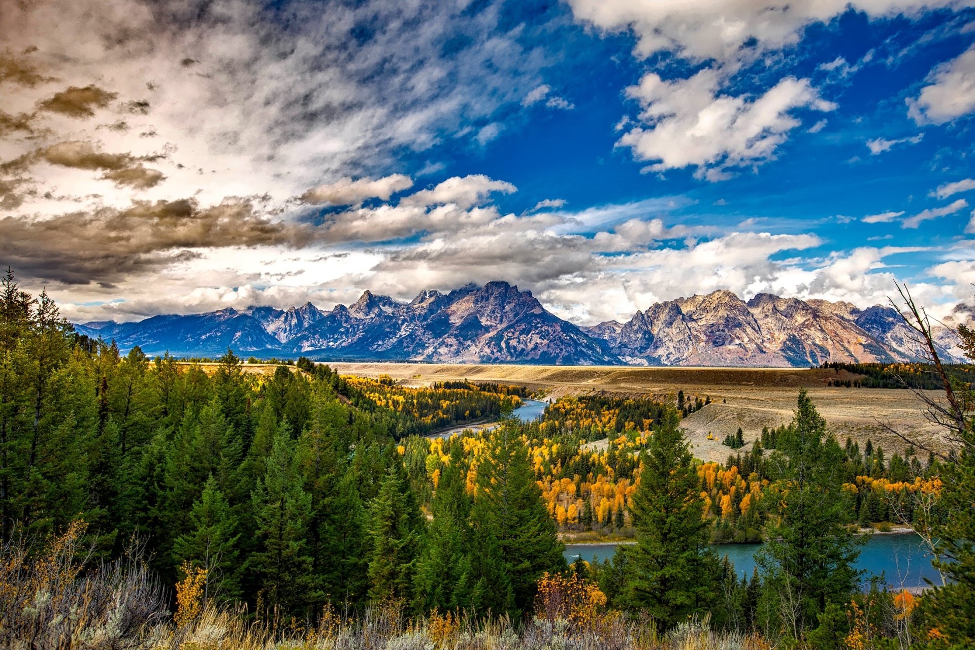 Vibrant fall landscape of Grand Teton National Park, Wyoming, featuring colorful trees, a winding river, forested foreground, and dramatic mountain peaks under a partly cloudy sky.