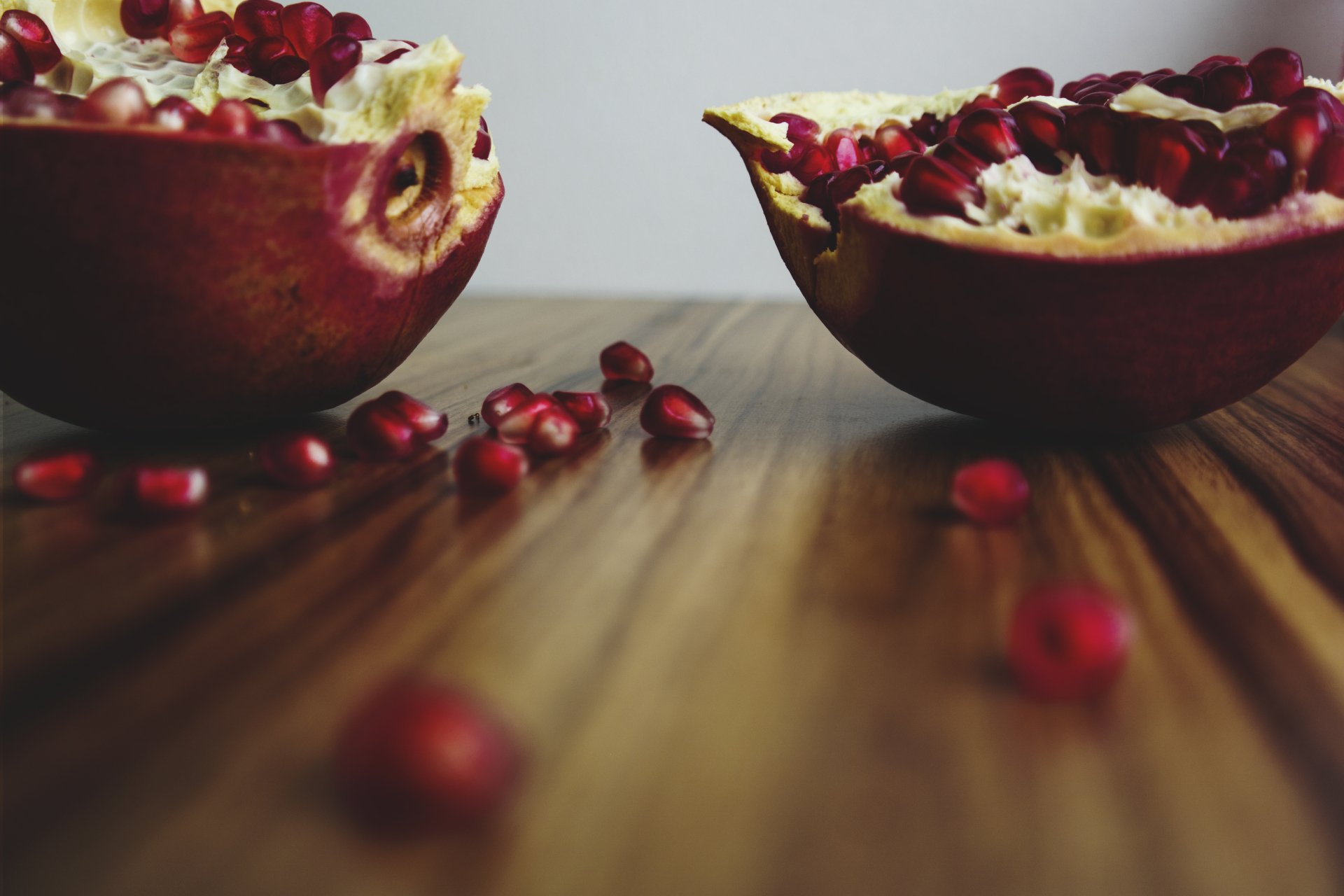 Halved pomegranate with scattered seeds on a wooden table — fruit, food; 5K Ultra HD PC desktop wallpaper and background.