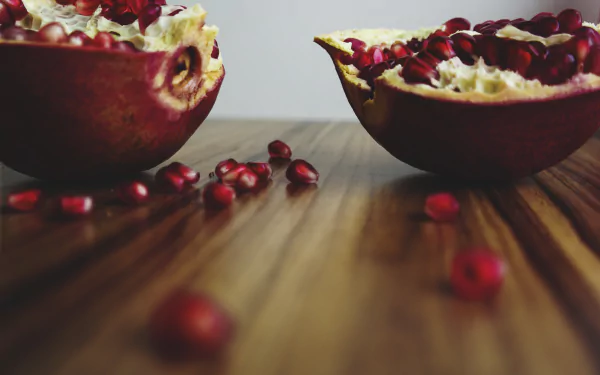 Halved pomegranate with scattered seeds on a wooden table — fruit, food; 5K Ultra HD PC desktop wallpaper and background.