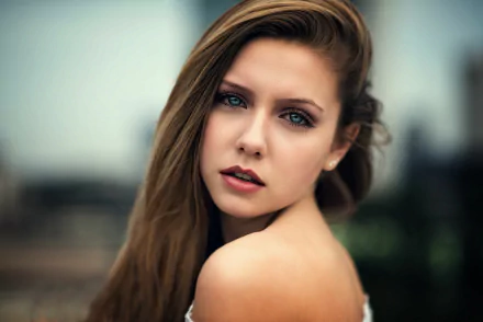 HD wallpaper of a brunette model with blue eyes. The woman is looking at the camera with an intense, captivating expression. The background is softly blurred, emphasizing her striking facial features.