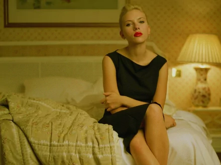 Scarlett Johansson sits elegantly on a bed, wearing a stylish black dress and bold red lipstick, illuminated by soft lighting in a cozy room. A striking HD wallpaper.