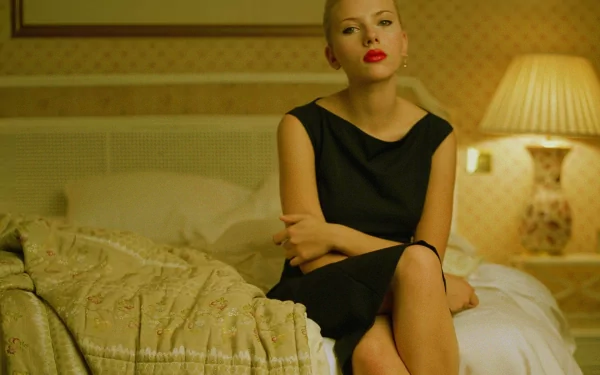 Scarlett Johansson sits elegantly on a bed, wearing a stylish black dress and bold red lipstick, illuminated by soft lighting in a cozy room. A striking HD wallpaper.