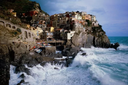 HD desktop wallpaper of Manarola, a picturesque man-made village perched on a rocky coastline with colorful buildings, waves crashing against the rocks, and a dramatic sky overhead.