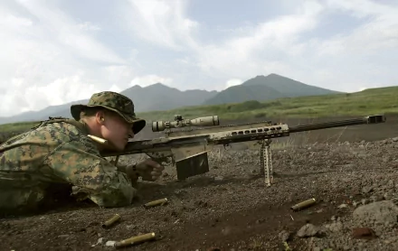 A soldier in camouflage lies prone on the ground, aiming a Barrett M82 .50 cal sniper rifle in a military setting, with mountains visible in the background under a partly cloudy sky. HD desktop wallpaper and background.