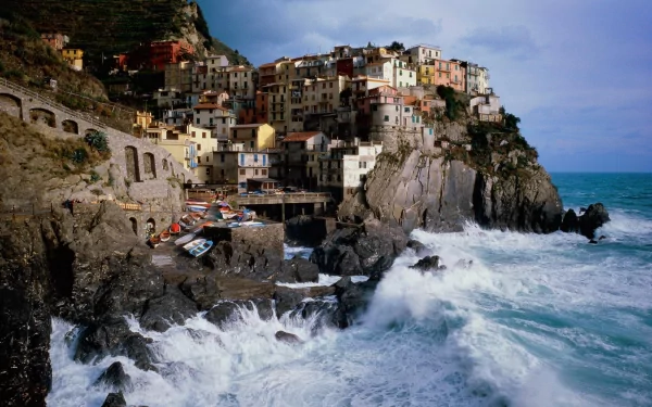 HD desktop wallpaper of Manarola, a picturesque man-made village perched on a rocky coastline with colorful buildings, waves crashing against the rocks, and a dramatic sky overhead.