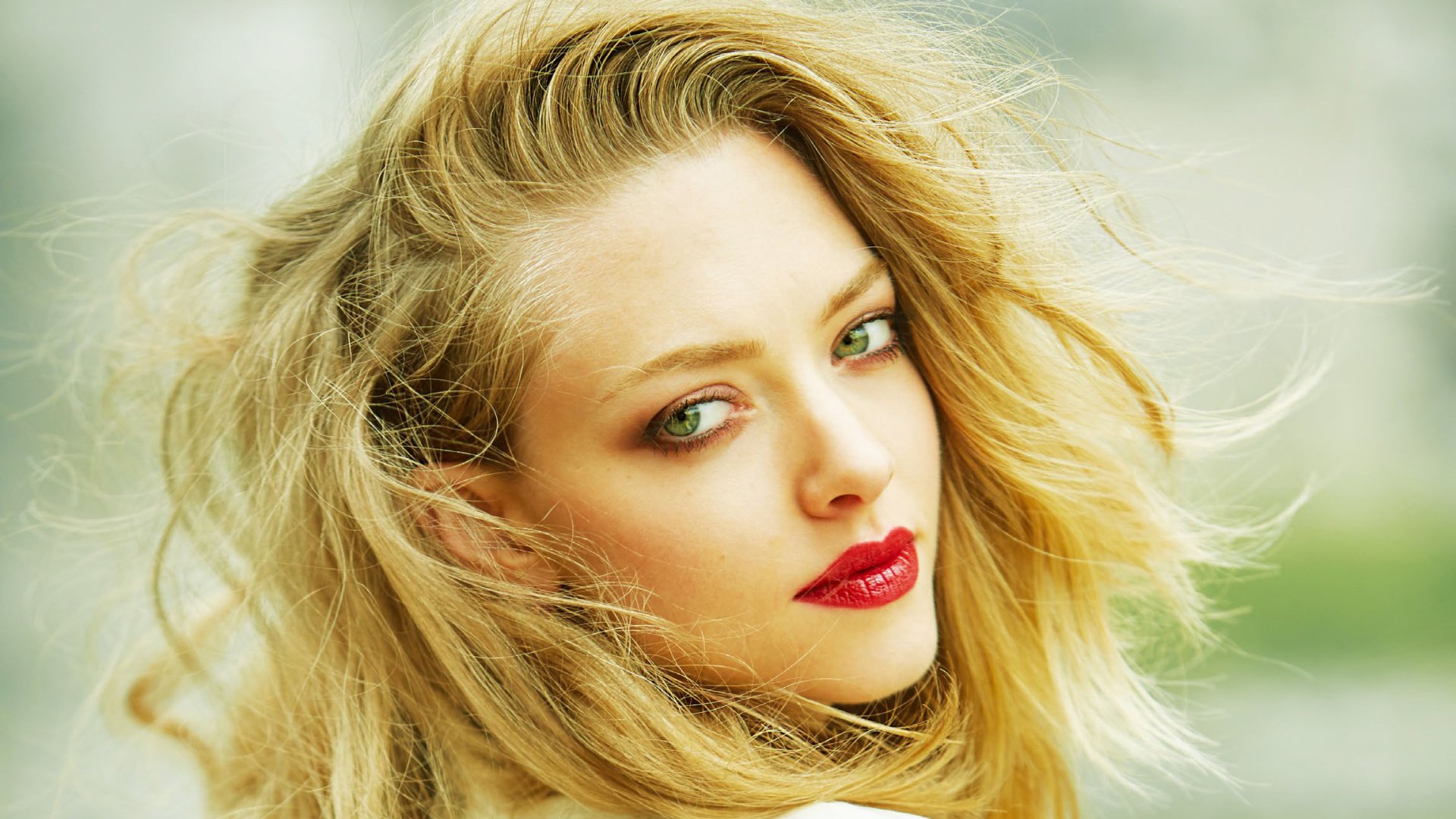 Close-up HD PC desktop wallpaper of a blonde woman’s face with green eyes, red lipstick and windblown hair set against a soft, blurred background.