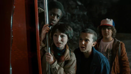 A scene from the TV show Stranger Things, featuring characters Lucas Sinclair, Mike Wheeler, Eleven, and Dustin Henderson. The young friends look worriedly at something off-screen.
