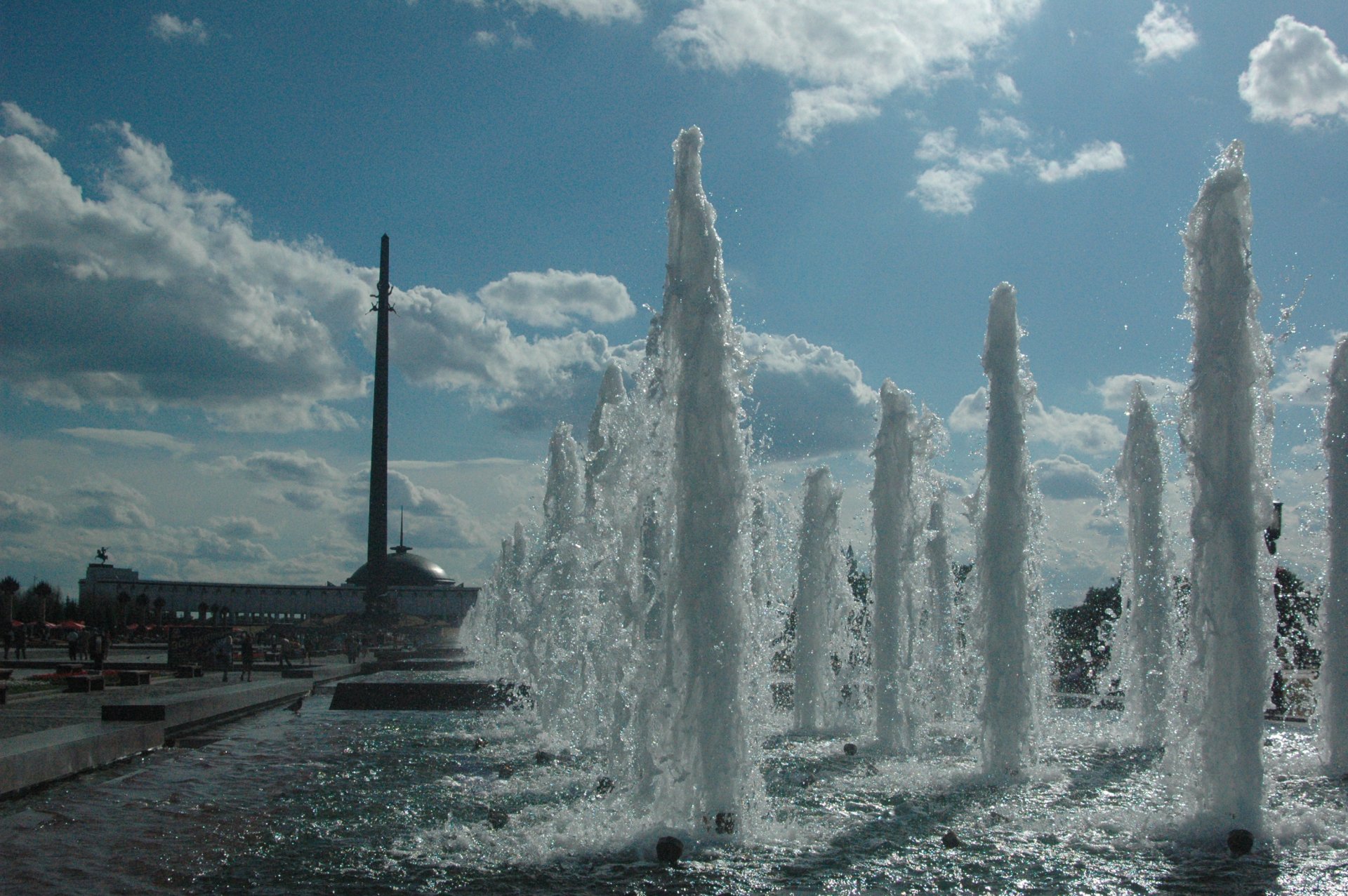 A dramatic view of a man-made fountain with towering water jets, set against a backdrop of clouds and sunlight, creating a serene and captivating scene for a HD desktop wallpaper.