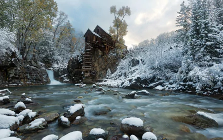 Stone Crystal Mill stands beside a flowing river surrounded by snow-covered trees in a serene winter landscape, captured in an HD desktop wallpaper.