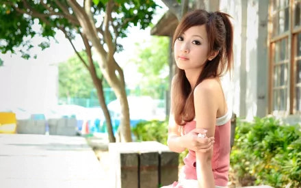 HD desktop wallpaper featuring a sensual Asian woman in a pink top, sitting outdoors with greenery and sunlight in the background.
