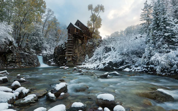 Stone Crystal Mill stands beside a flowing river surrounded by snow-covered trees in a serene winter landscape, captured in an HD desktop wallpaper.
