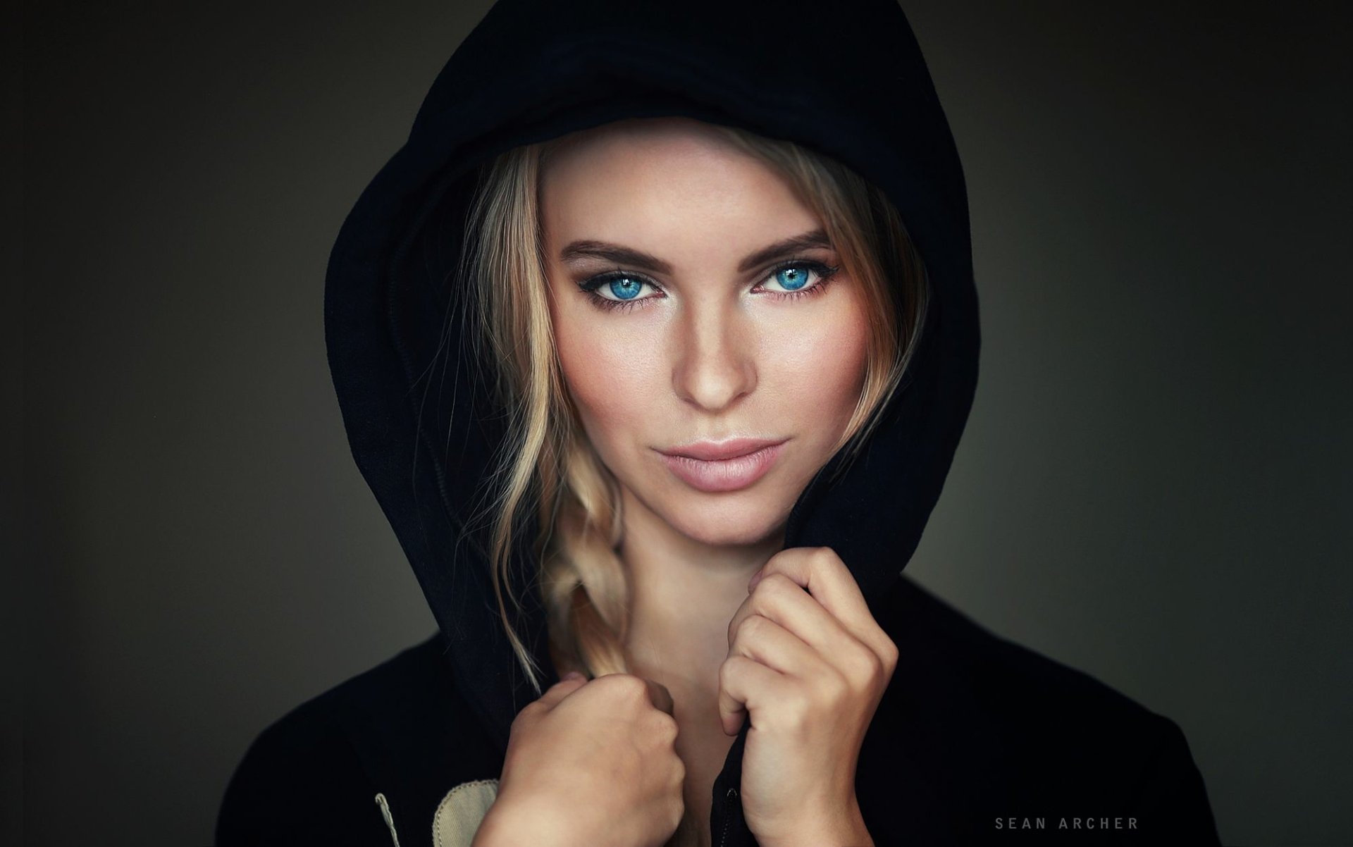 Close-up HD portrait of a blonde woman with blue eyes and a braid, wearing a black hooded top, set against a dark background.